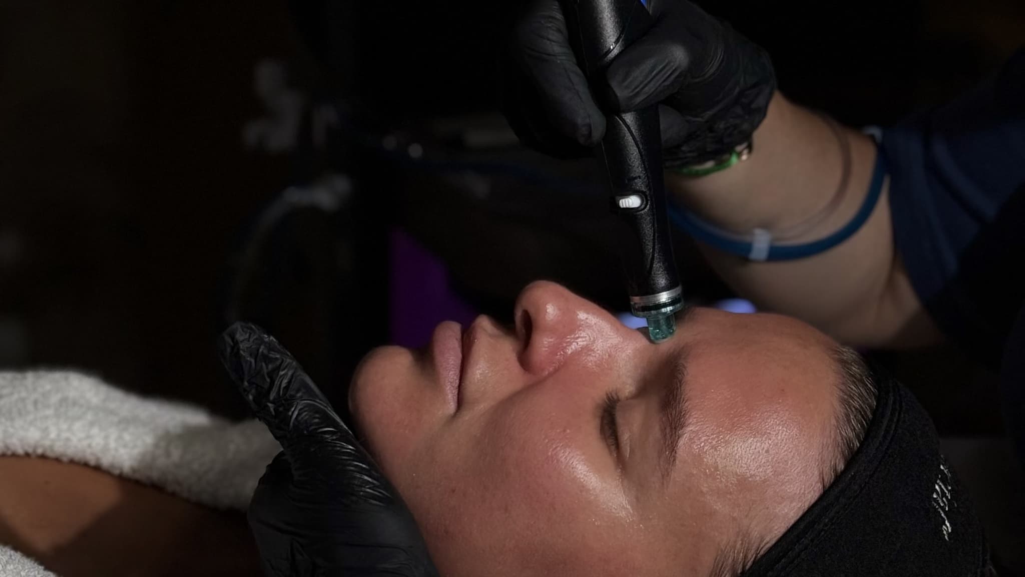 A gloved therapist holds a HydraFacial handpiece against the side of a client's nose as she lies back with her eyes closed, a black headband holding back her hair