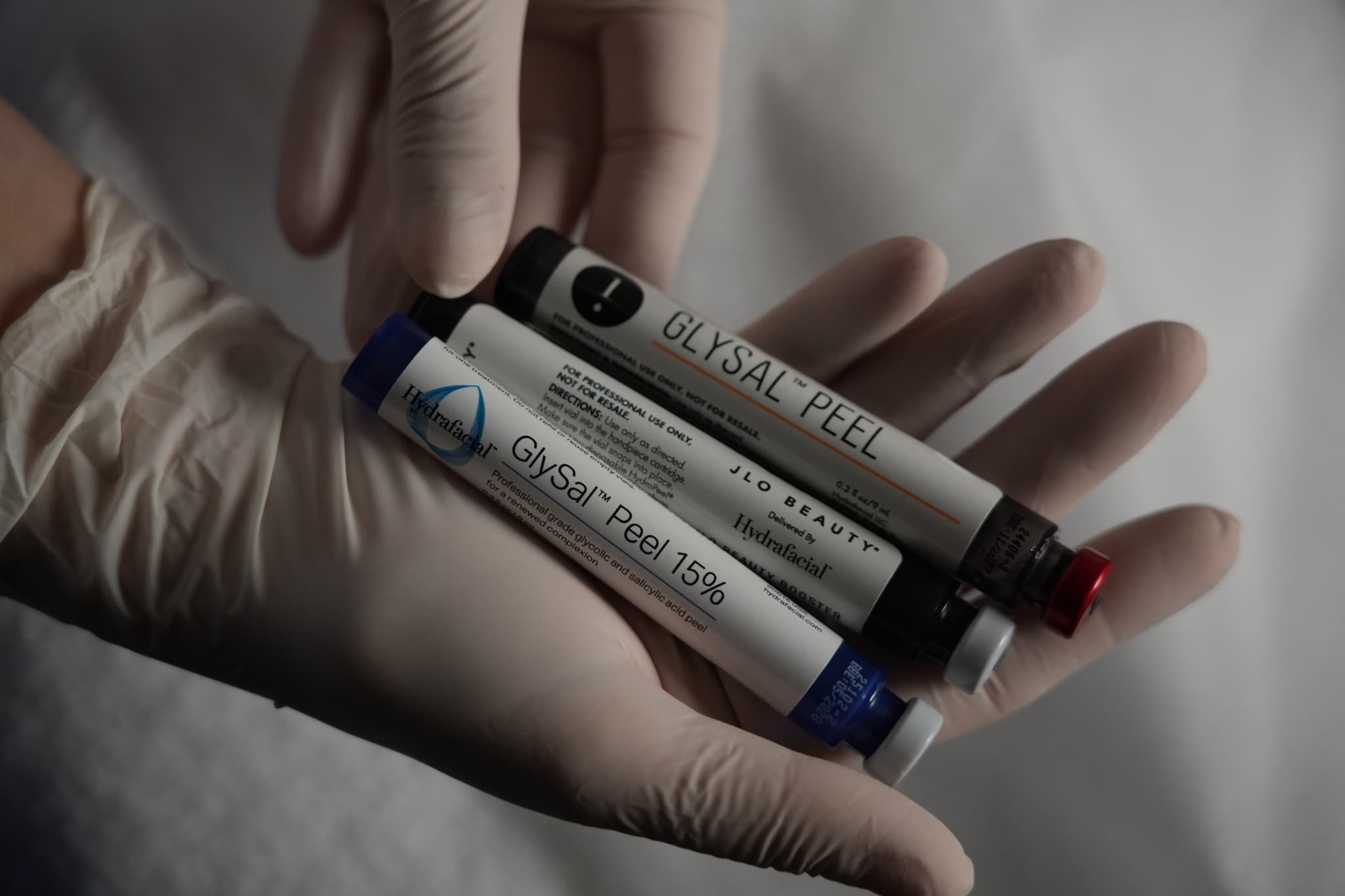 A gloved hand holding three HydraFacial GlySal Peel vials, one labelled GlySal Peel 15%, against white fabric