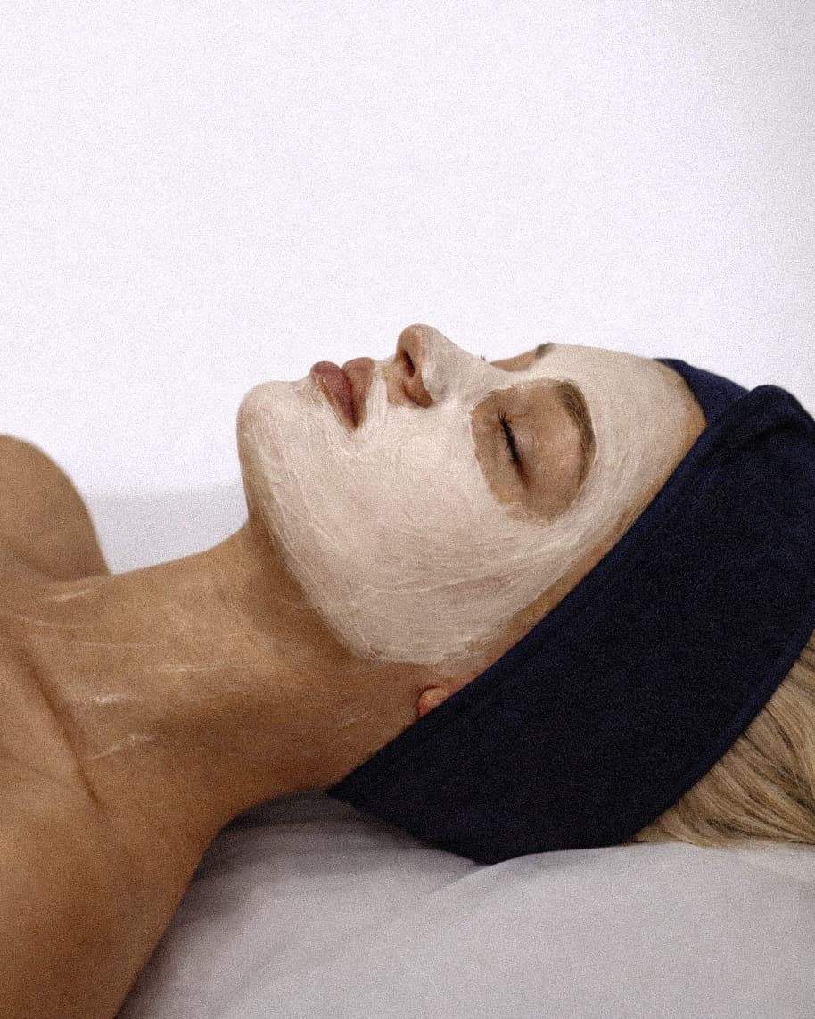 A woman lying back on a treatment bed with her eyes closed, a thick white cream mask covering her face and a navy towelling headband holding her hair off her forehead