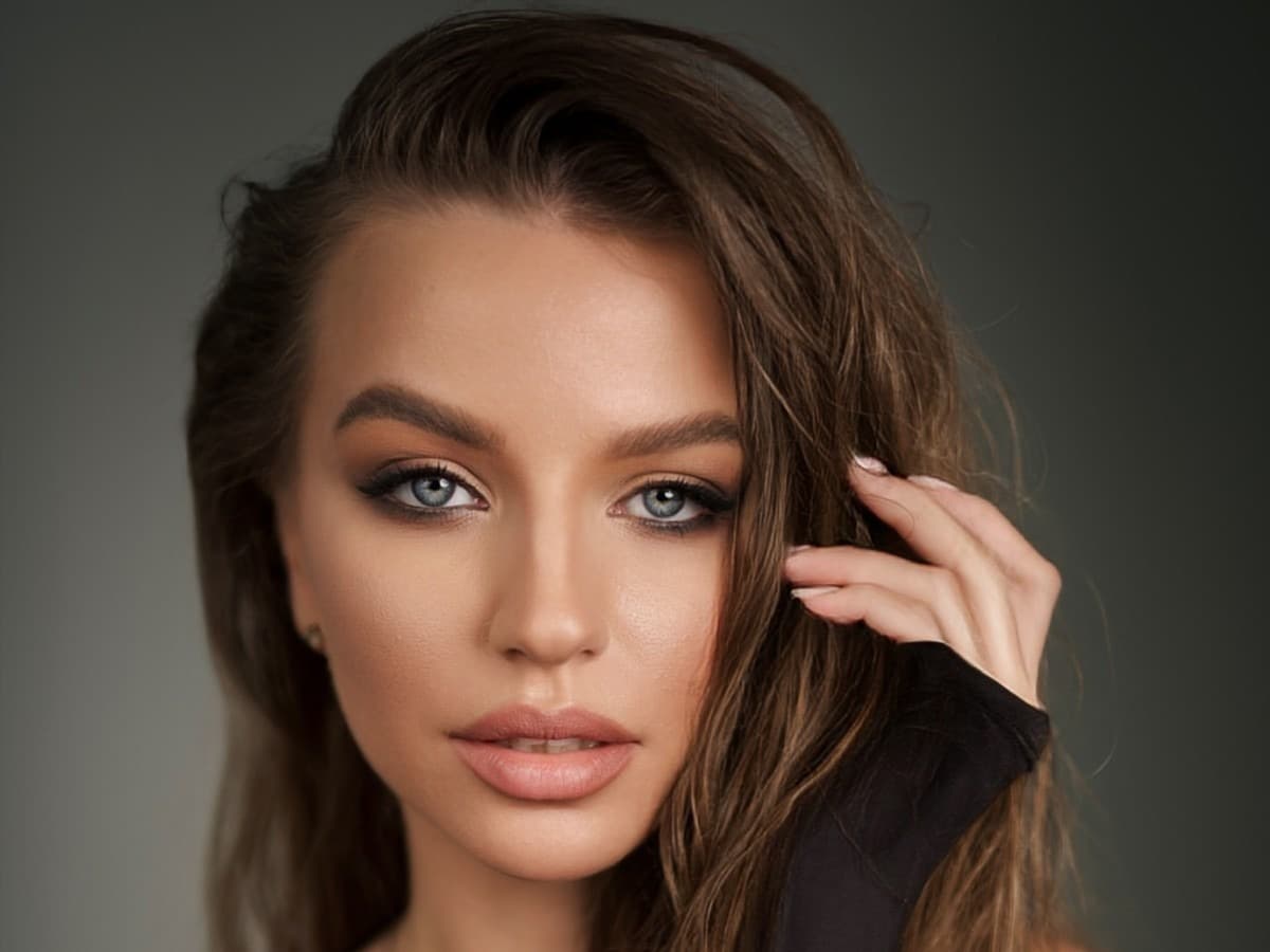 A woman with long brown hair in loose, textured waves, one hand lifting a section away from her face