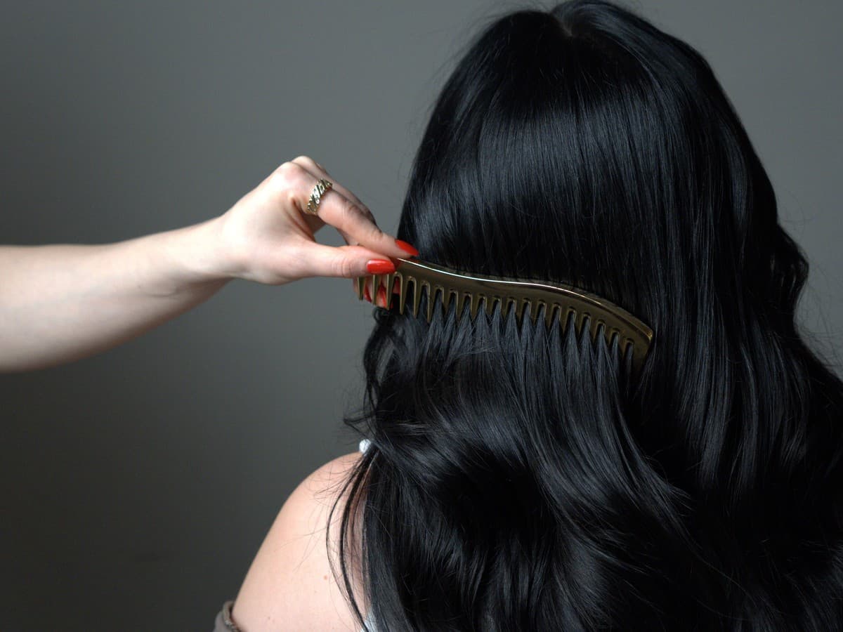 A gold wide-tooth comb drawn through long dark wavy hair, seen from behind