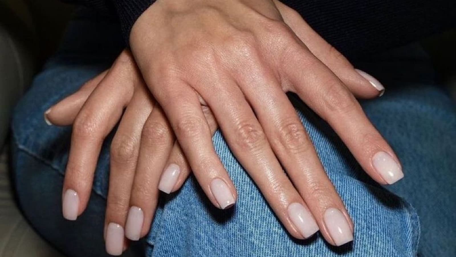 Short square nails in a sheer milky pink finish, hands crossed over a knee in blue jeans